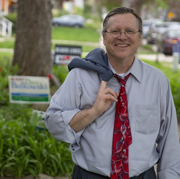 Dave Bulkowski smiling outdoors on a Grand Rapids street, a sport coat slung over his shoulder, with one of his campaign yard signs behind him.